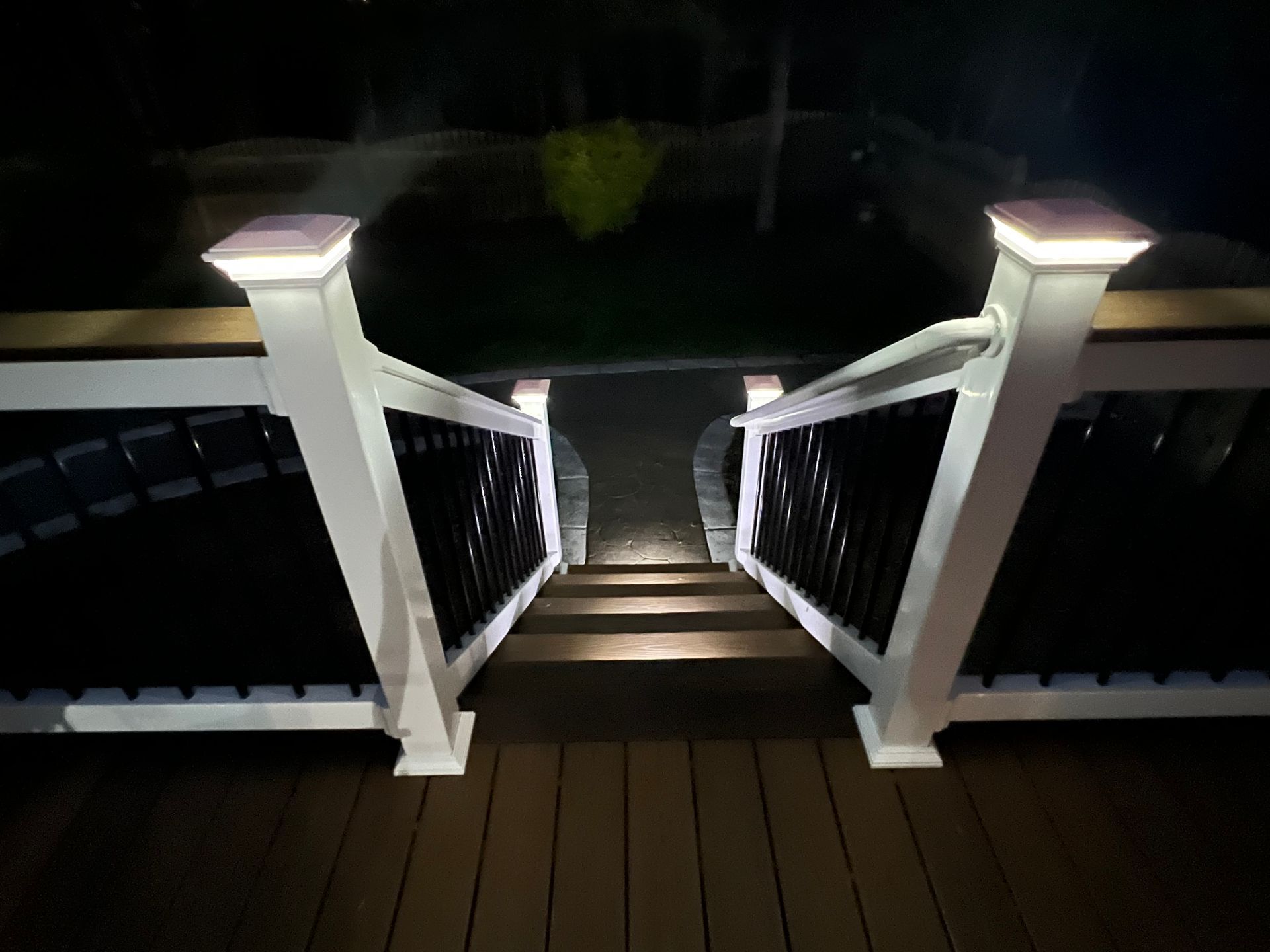 Nighttime view down a lit wooden staircase with white railings leading to a dark yard.