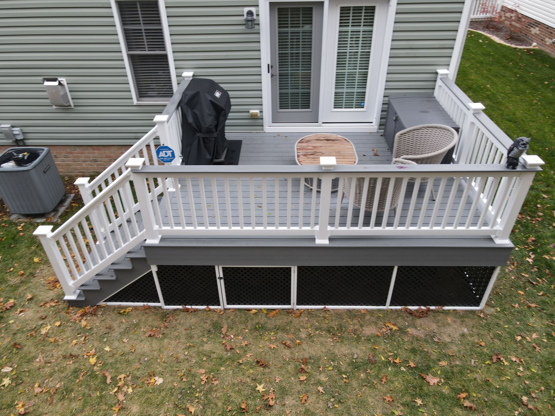 Small gray deck with white railing, stairs, grill, and chairs beside a house.