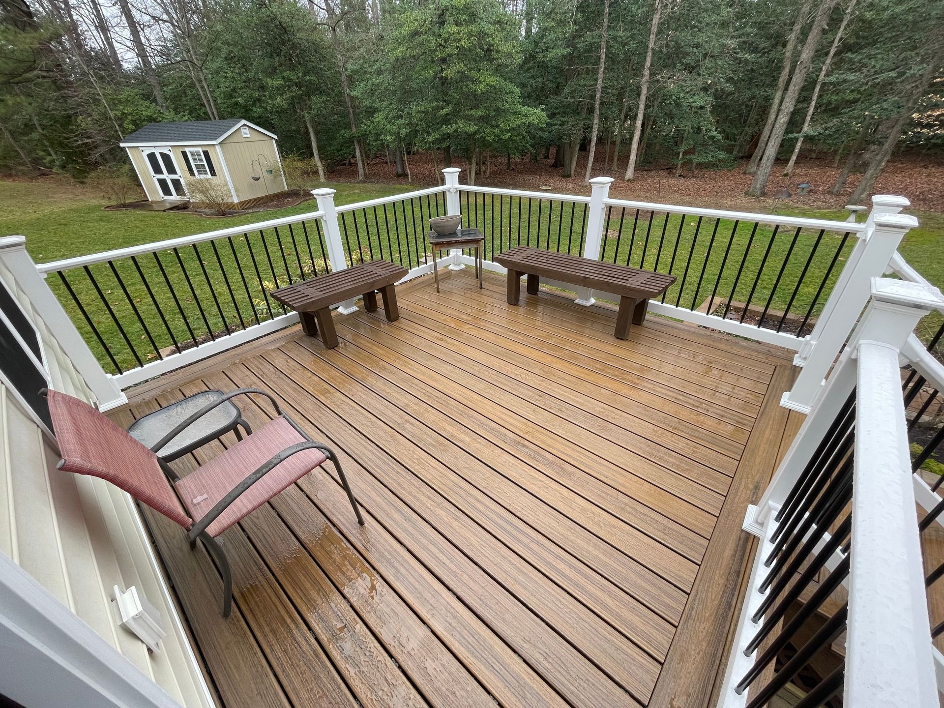 Wooden deck with benches and chairs overlooking a wooded yard and small shed