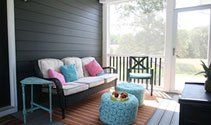 Cozy porch seating area with a gray loveseat, colorful pillows, turquoise table, and bright window view