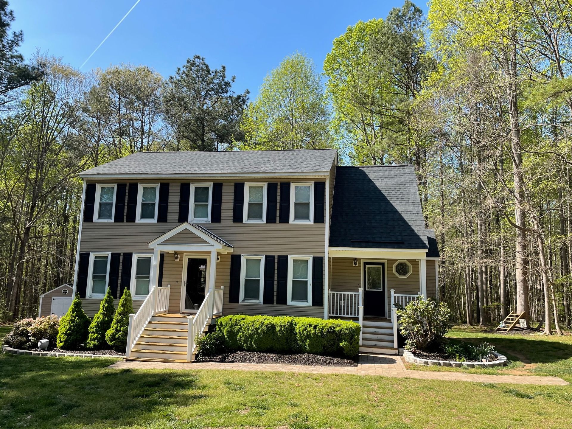 Two-story beige house with black shutters and a dark roof, surrounded by trees and a lawn in daylight