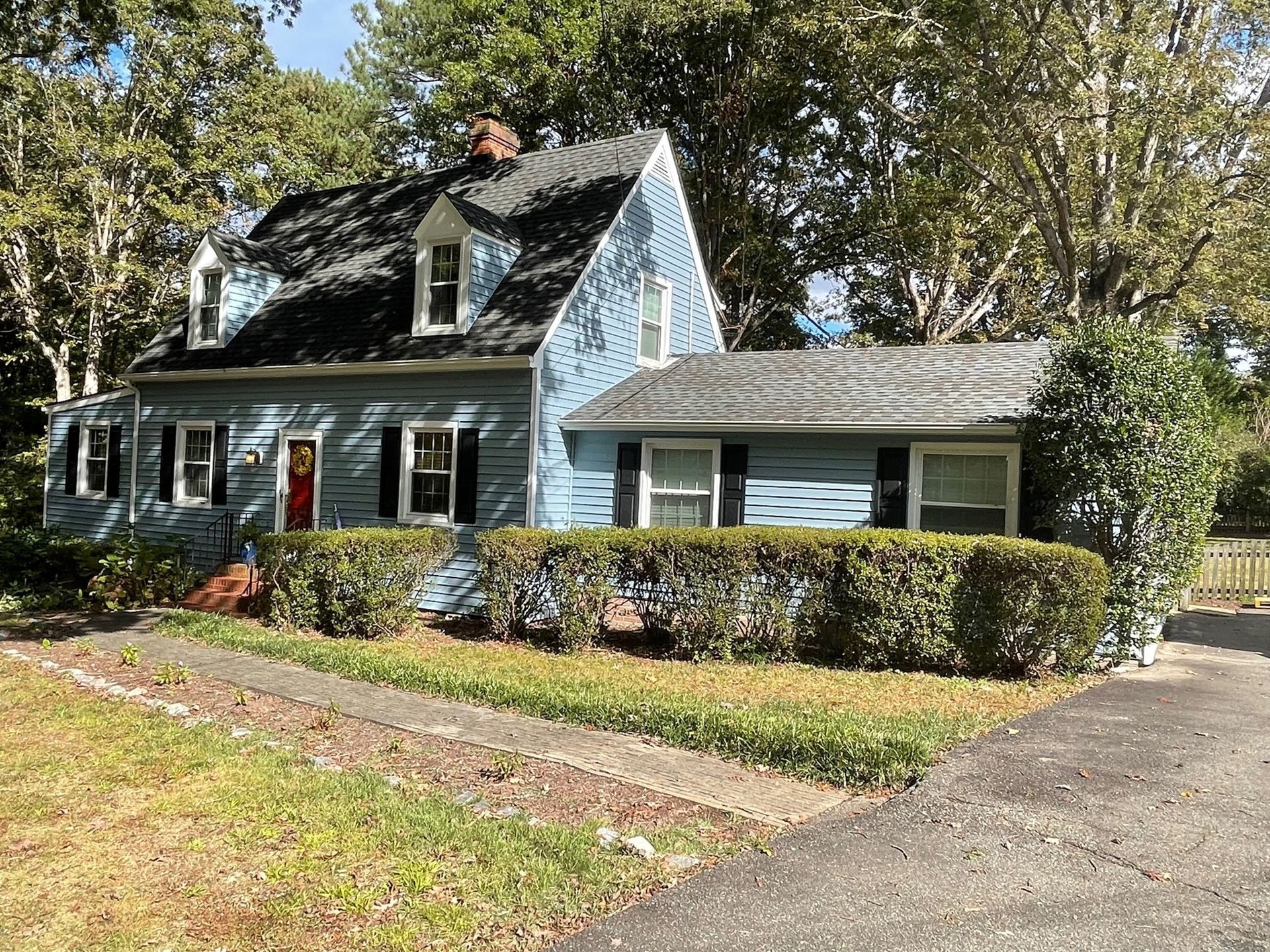 Blue house with dormer windows, white trim, and a front hedge beside a driveway in a leafy yard