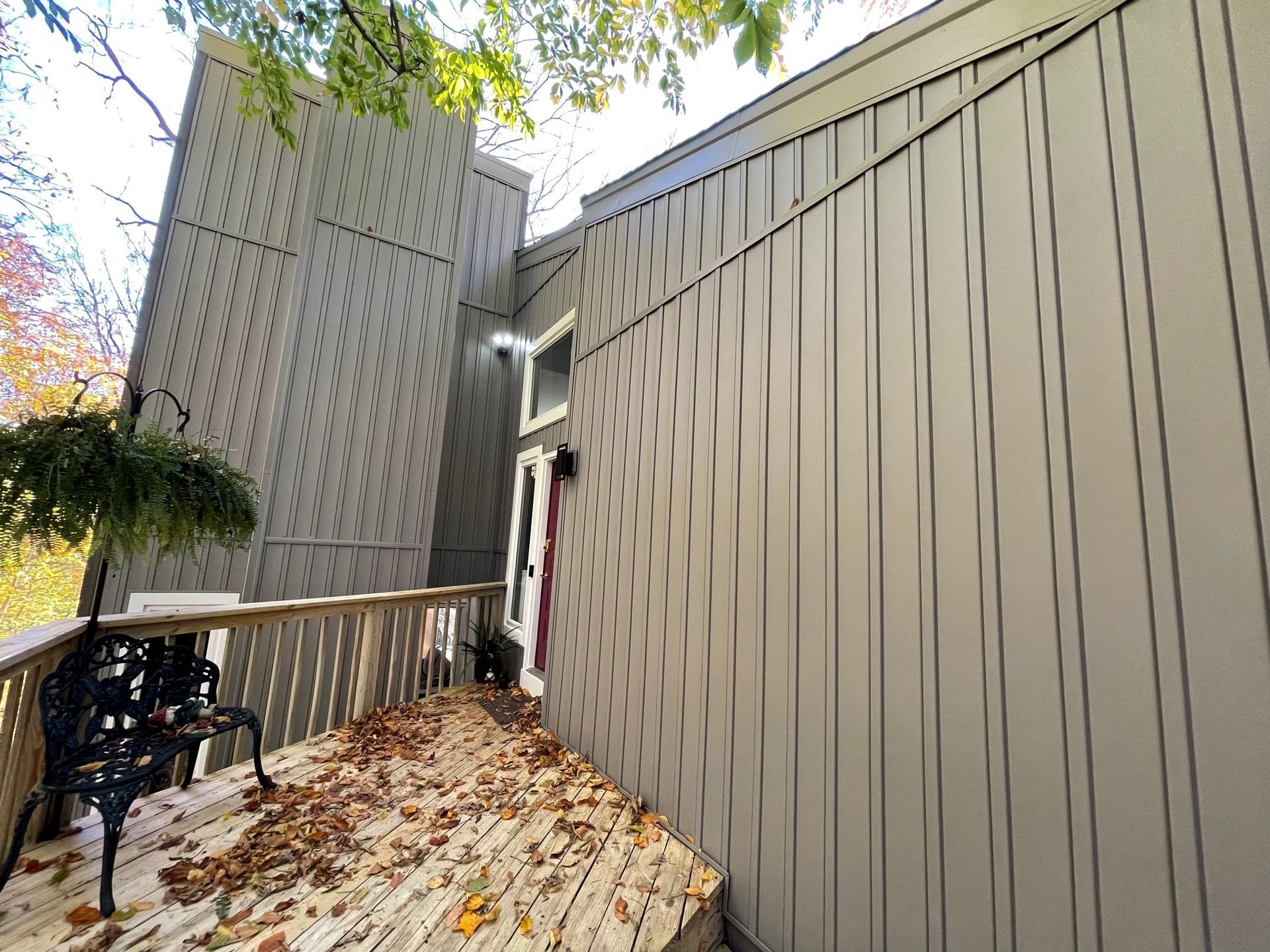 Wooden apartment walkway with fallen leaves, railing, and tan siding beside a tall building.