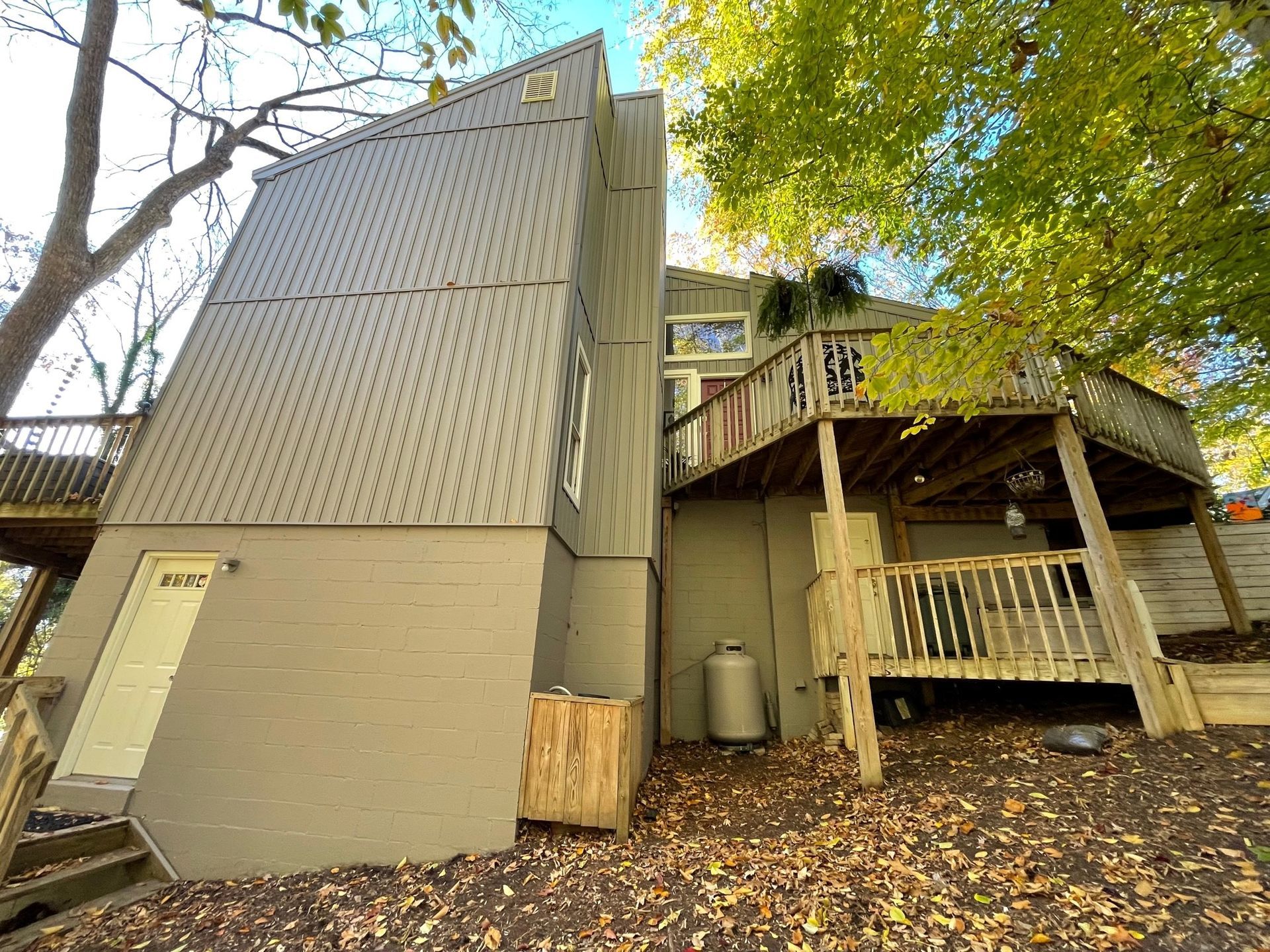 Tall beige building beside a wooden deck and stairs under leafy trees, covered in fallen leaves
