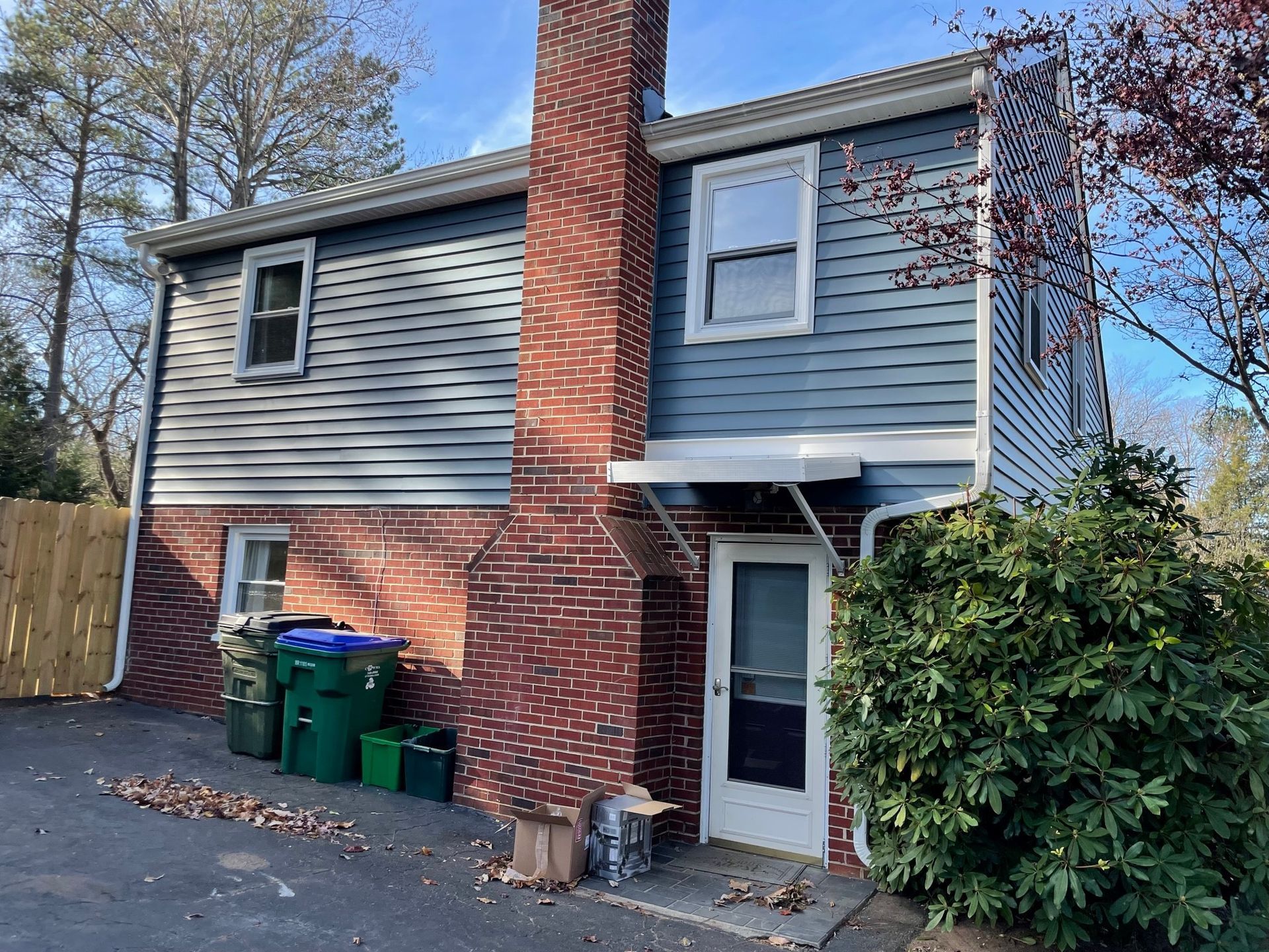 Back of a brick house with blue siding, white door, chimney, trash bins, and a large bush