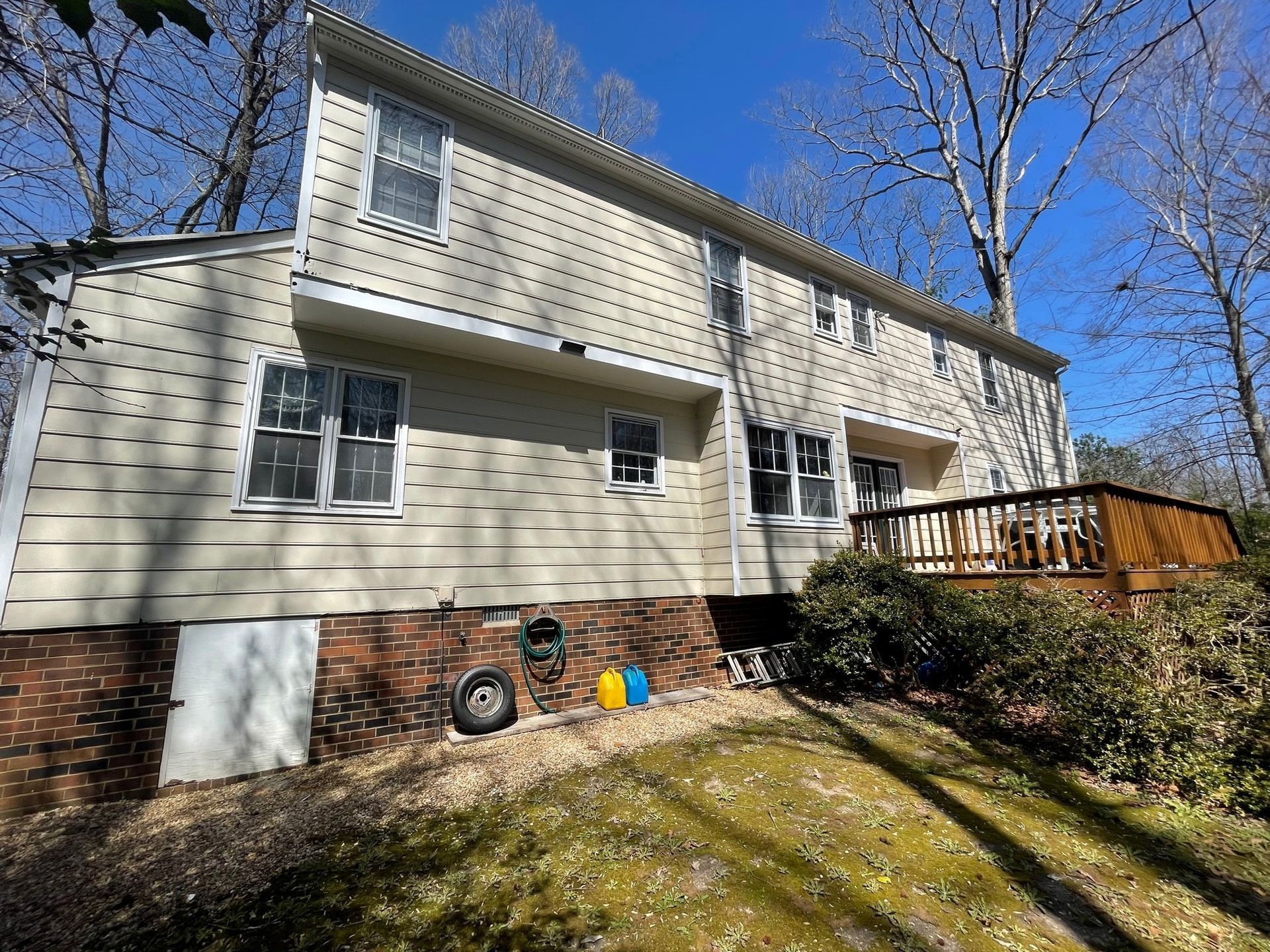 Two-story beige house with brick foundation, wooden deck, and leafless trees under a clear blue sky.