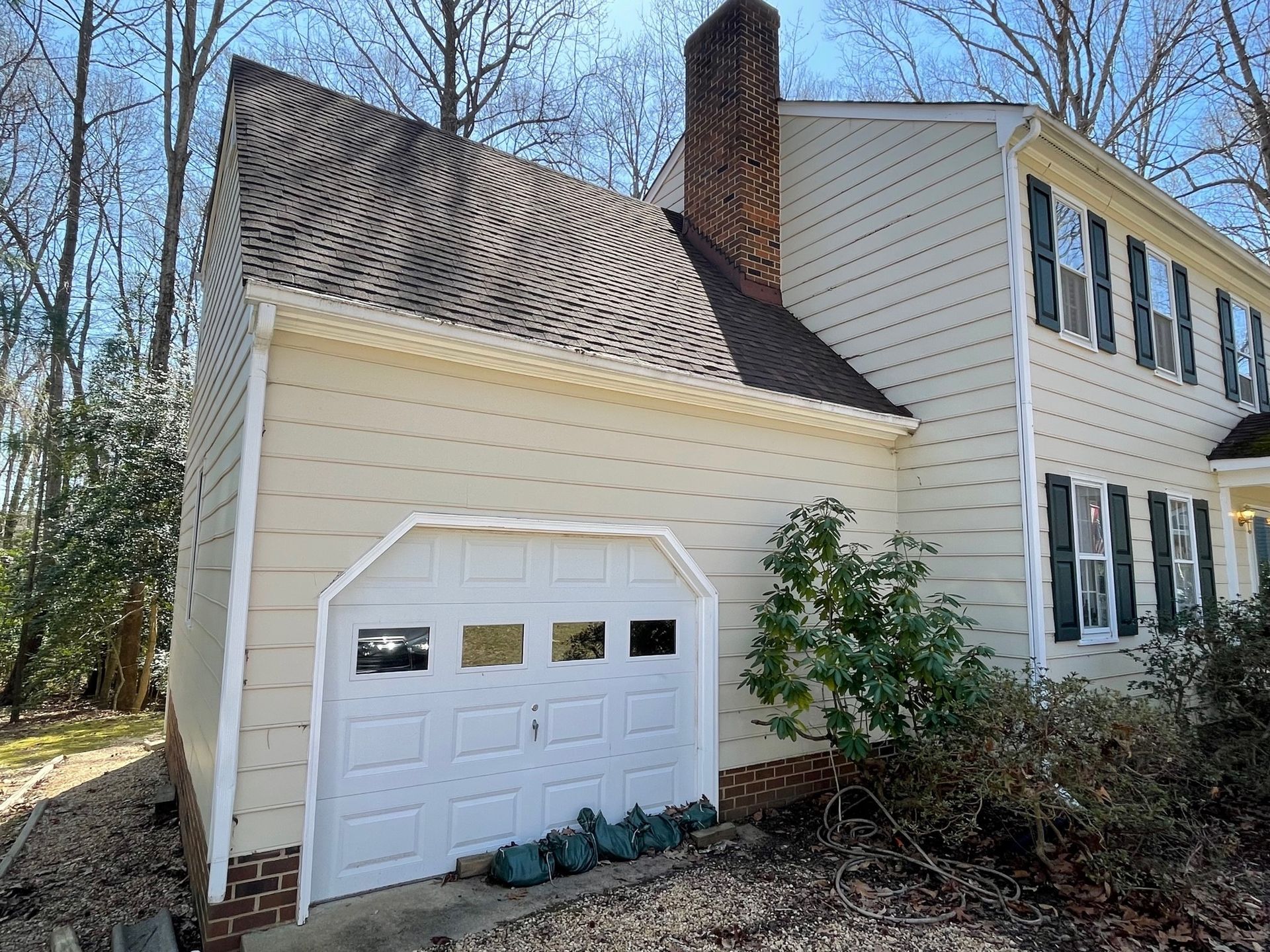 Cream-sided house with a garage, dark shingled roof, and tall brick chimney in a wooded yard.