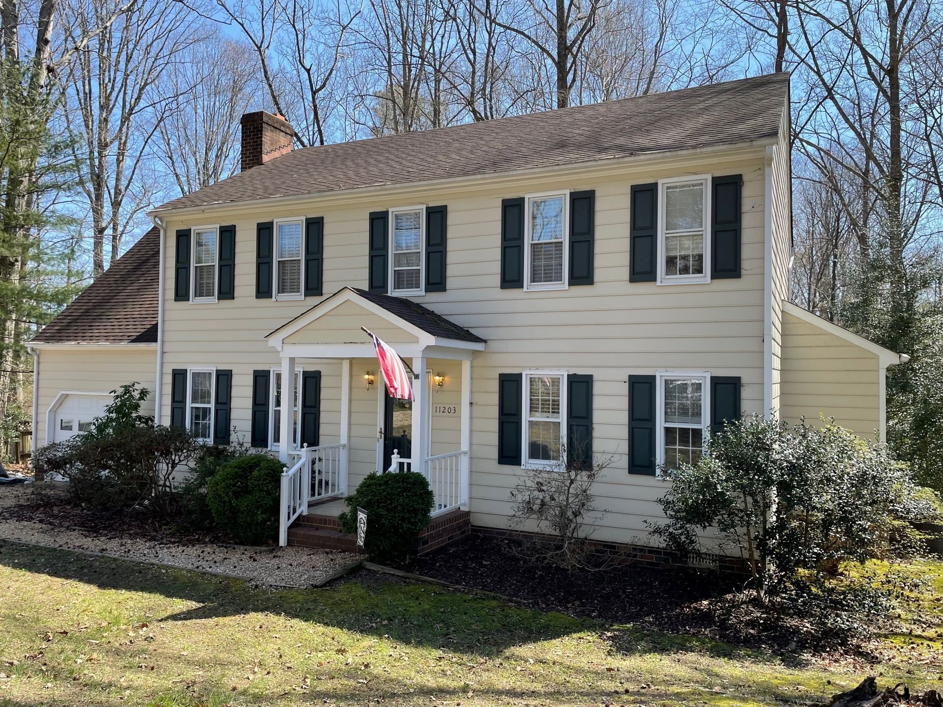 Two-story beige house with black shutters, white porch, and an American flag in a wooded yard