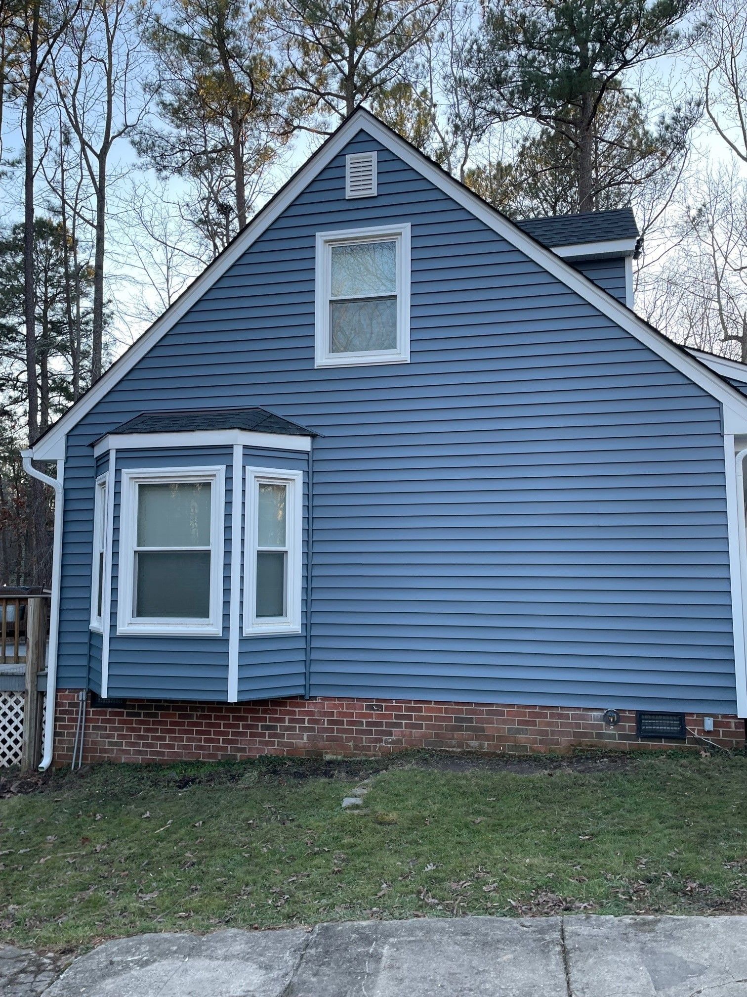 Blue house with white trim, brick foundation, and a small bay window in a wooded yard
