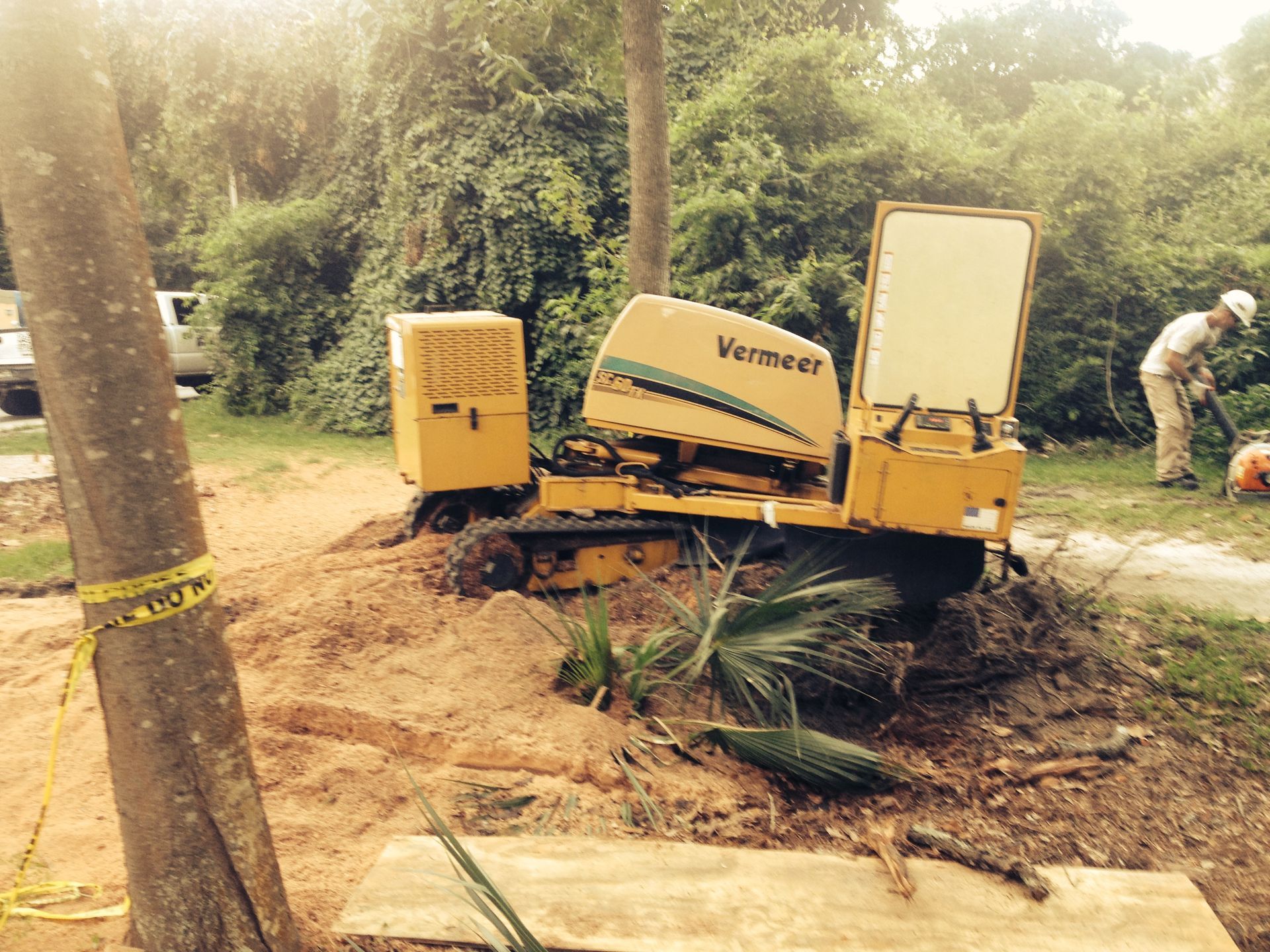 Yellow Vermeer stump grinder grinding a tree stump. A worker is nearby with tools.