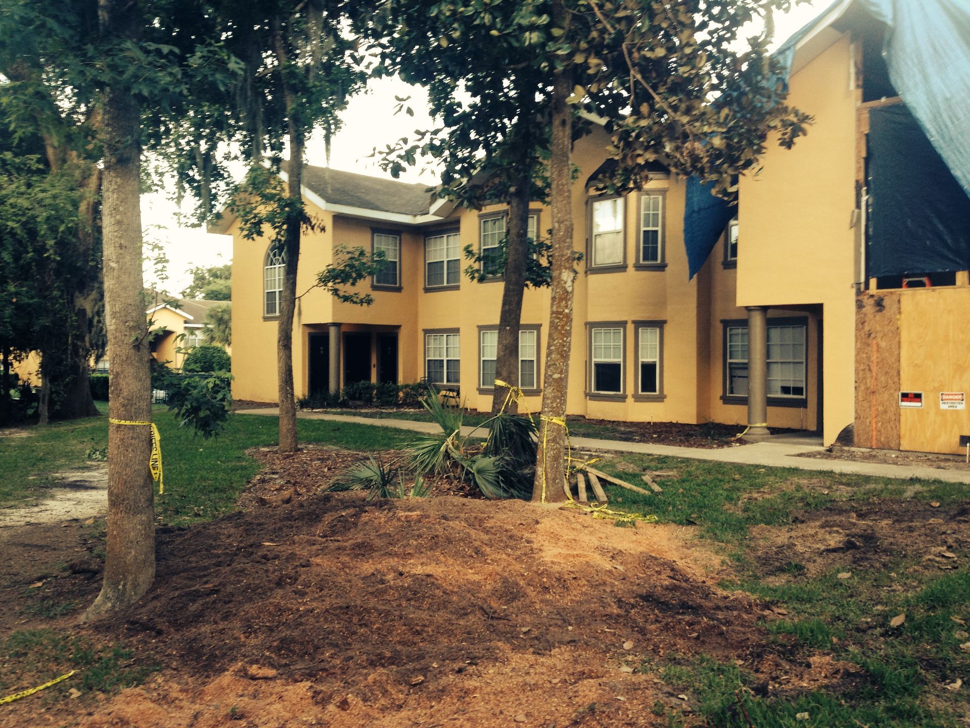 Two-story yellow buildings with trees and a dirt pile in front. Construction with blue tarp on the right.