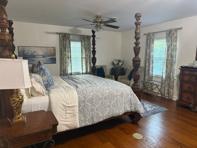 Bright bedroom with a four-poster bed, patterned bedding, wooden furniture, and windows with blinds and curtains