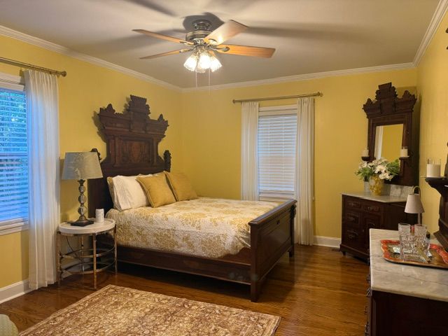 Warm yellow bedroom with ornate wood bed, dresser, mirror, and ceiling fan by windows.