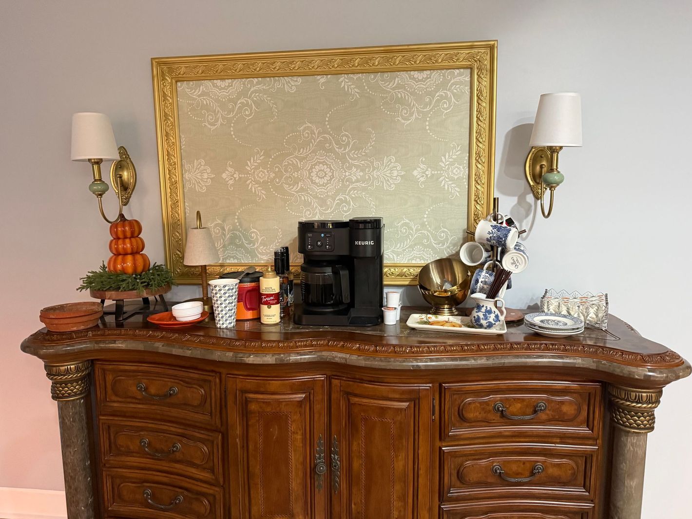 Wooden dresser with coffee maker, jars, and decorative items beneath a gold-framed mirror and wall sconces