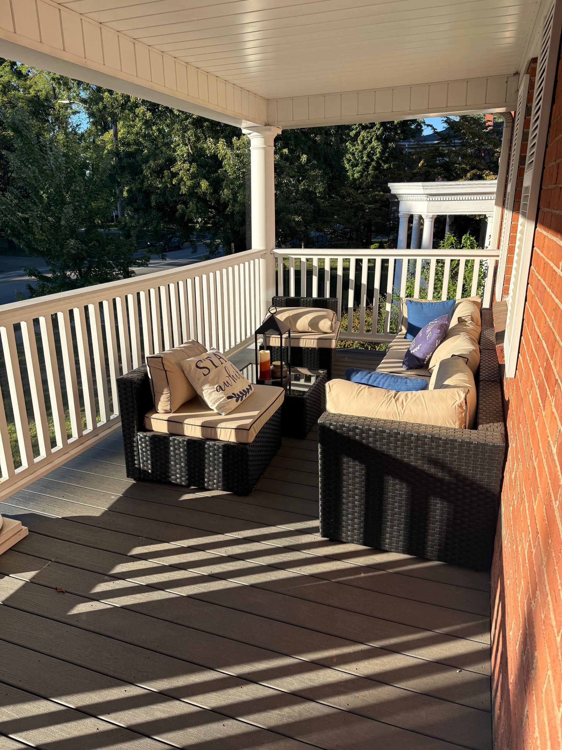 Sunlit porch with wicker chairs, coffee table, and striped cushions, overlooking a yard