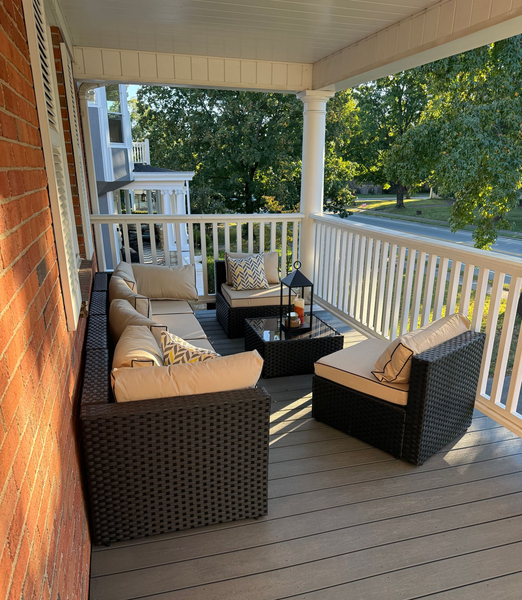 Covered porch with wicker patio furniture, beige cushions, and a view of green trees beyond the white railing.