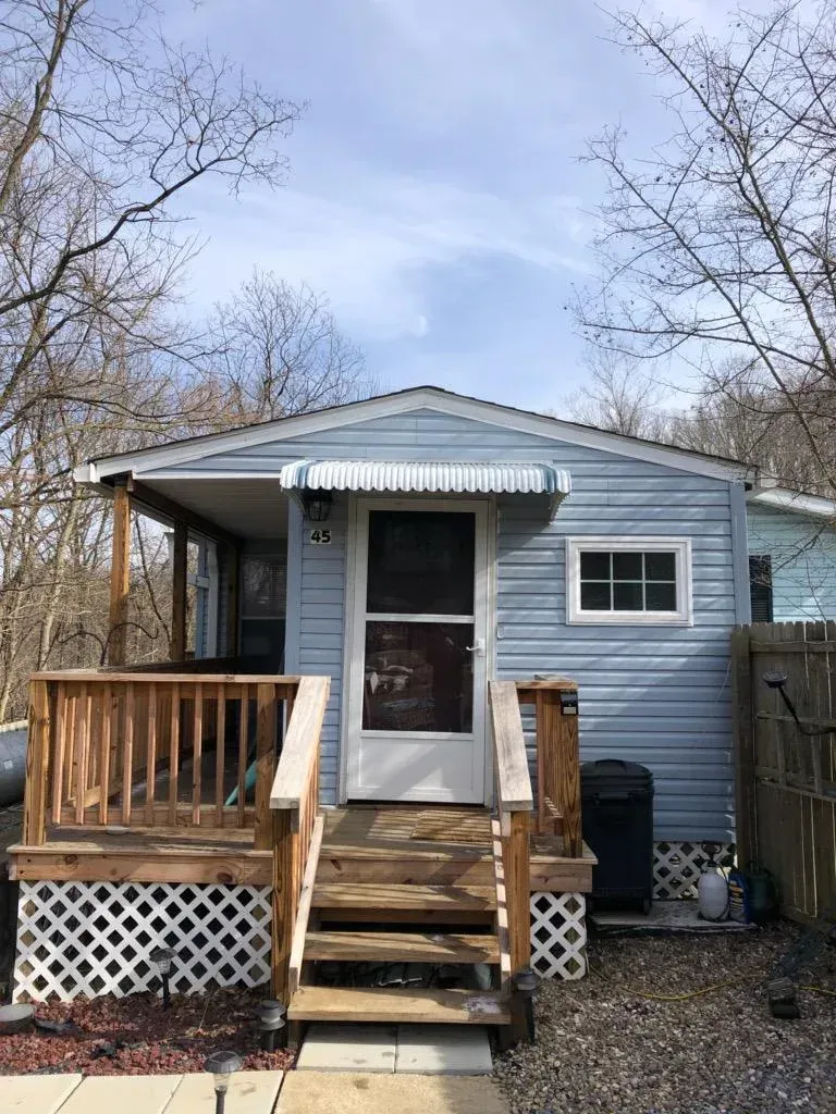 Blue siding cottage with porch and steps, awning over the door. Brown railing, lattice, and a small window.