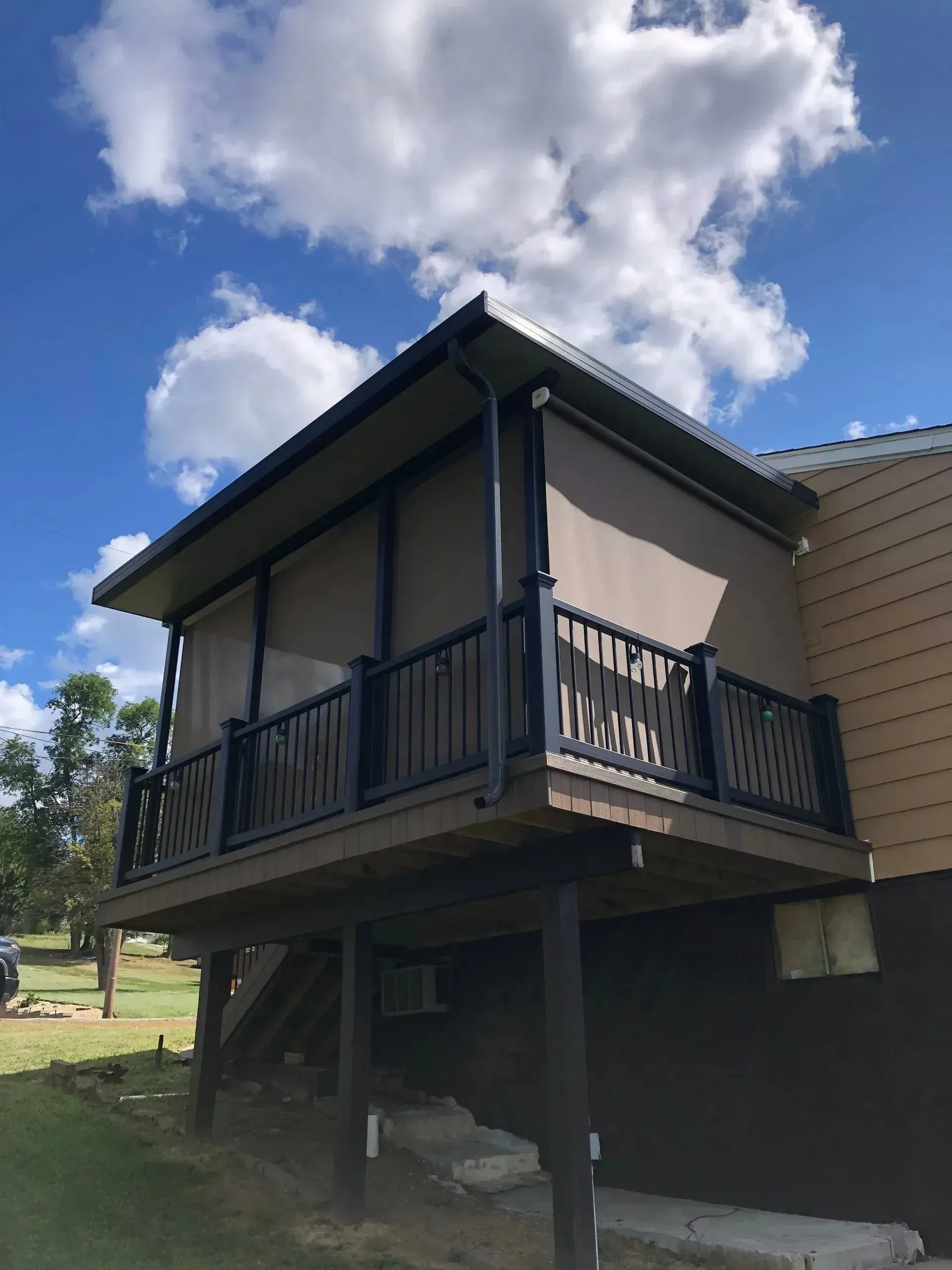 Covered deck with brown shades, black railing, and brown house siding under a partly cloudy blue sky.