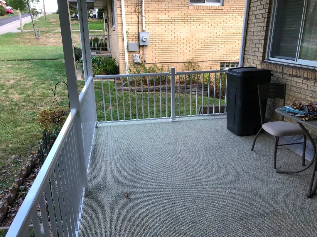 Covered porch with metal railing, carpeted floor, and view of a lawn. Black trash can, small table, and chair present.