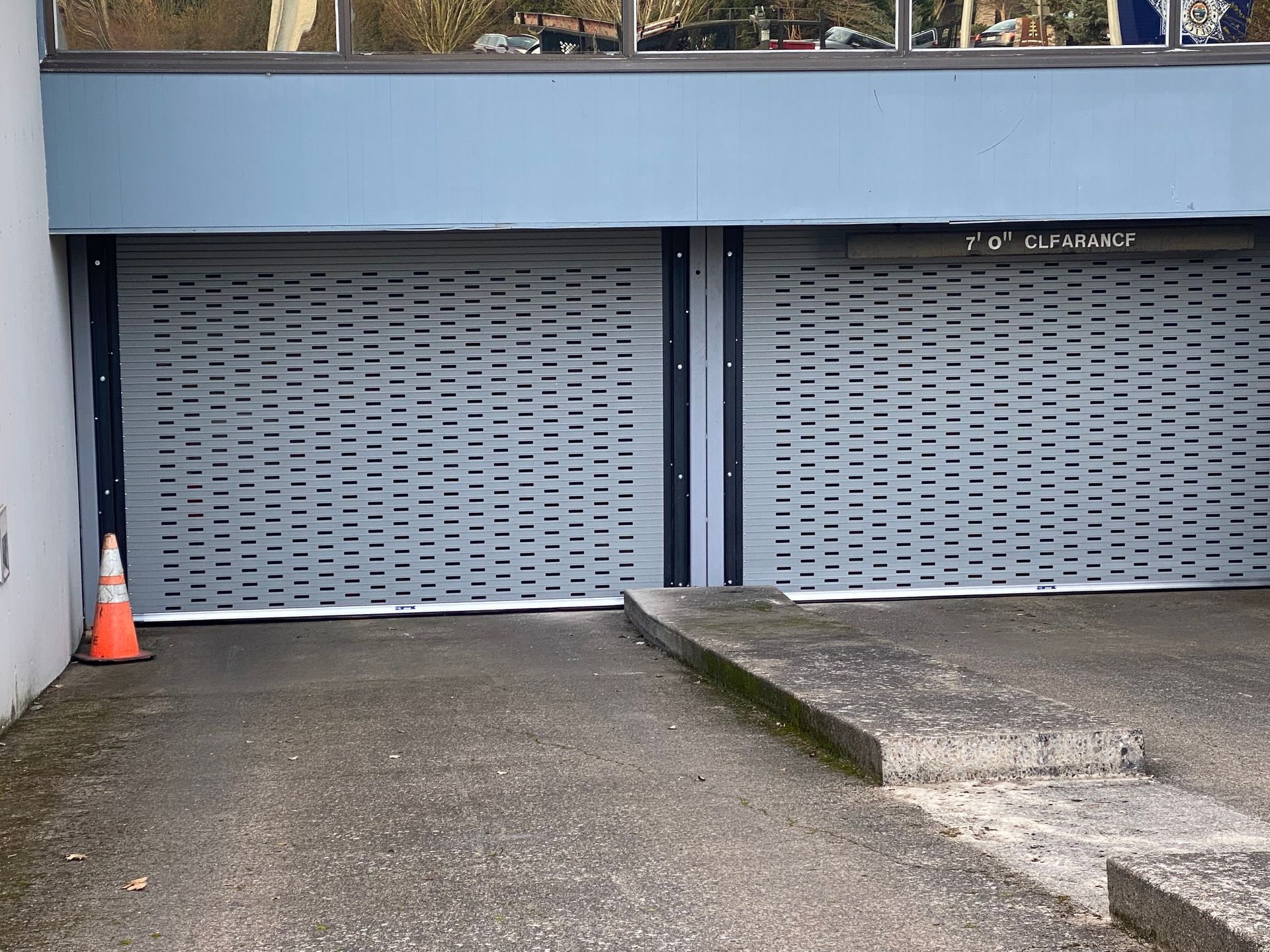 Two closed, gray, perforated roller shutters. Orange traffic cone on left. Concrete surface.