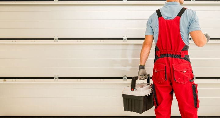 A man in red overalls is standing in front of a garage door holding a toolbox.