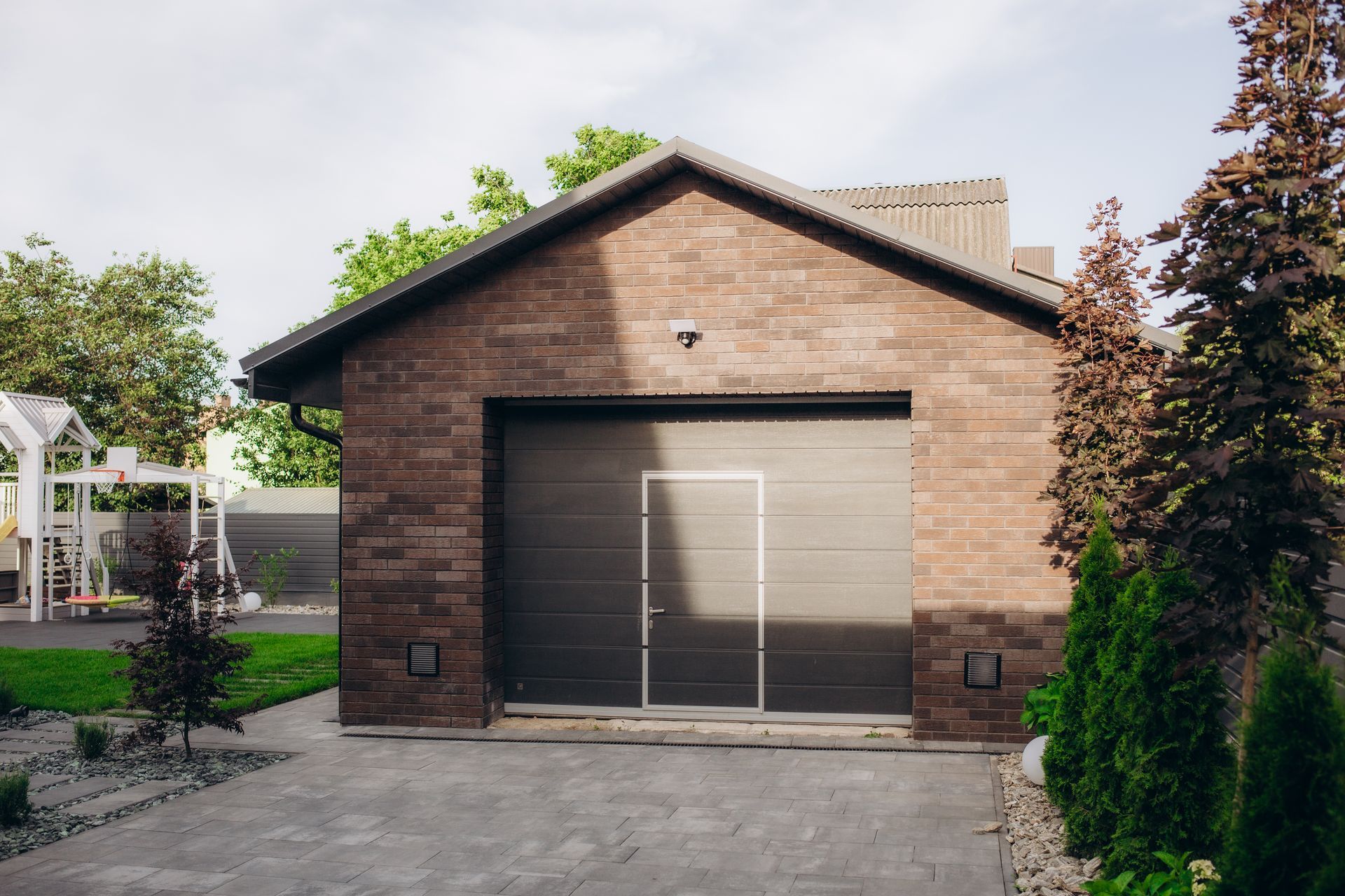 A brick garage with a black garage door is in the backyard of a house.