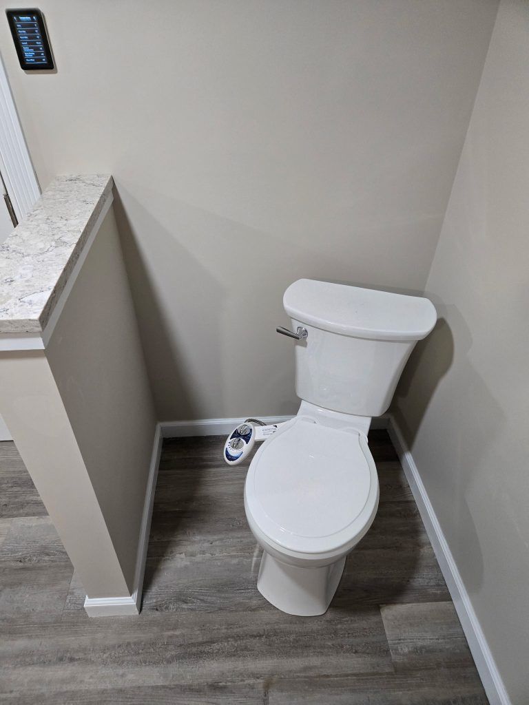 Small bathroom with a white toilet, gray wood-look floor, and a wall-mounted remote on the left.