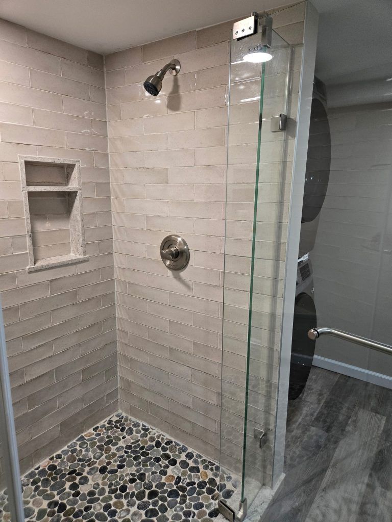 Glass shower with tiled walls, pebble floor, built-in niche, and chrome fixtures in a bathroom.