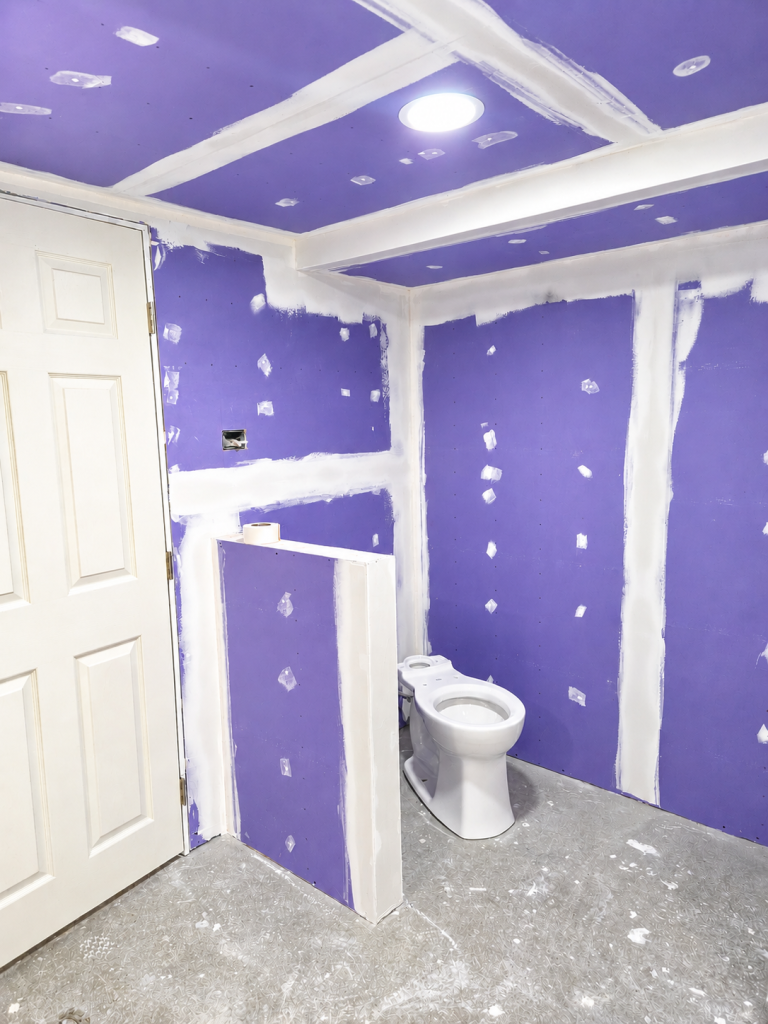 Small bathroom under renovation with purple drywall, white trim, toilet, and unfinished ceiling.