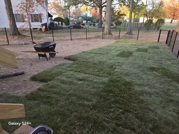 Freshly laid sod in a fenced yard with a wheelbarrow on a dirt path at dusk.