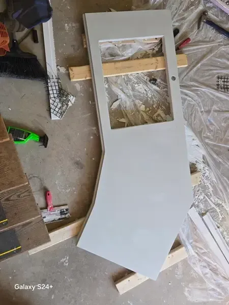 Gray painted wooden panel with a rectangular cutout, resting on a workshop floor with tools and plastic sheeting.