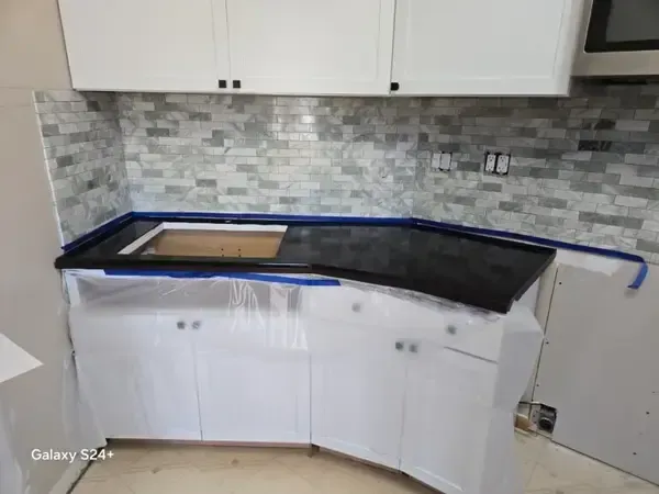 Partially renovated kitchen with white cabinets, gray tile backsplash, and a countertop wrapped in protective plastic