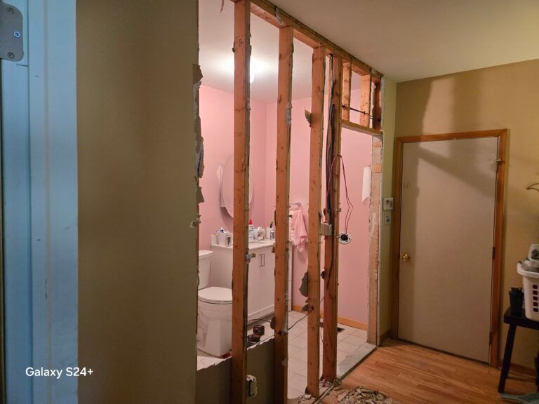 Partially demolished bathroom wall exposing studs and plumbing in a house interior