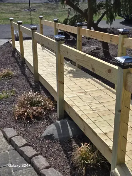 Wooden ramp with handrails over a gravel path, leading through a grassy outdoor area.