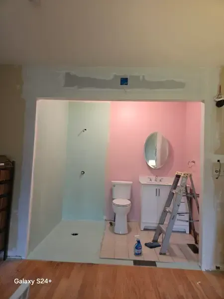 Bathroom under renovation with pink walls, toilet, sink, ladder, and shower area covered in protective plastic.