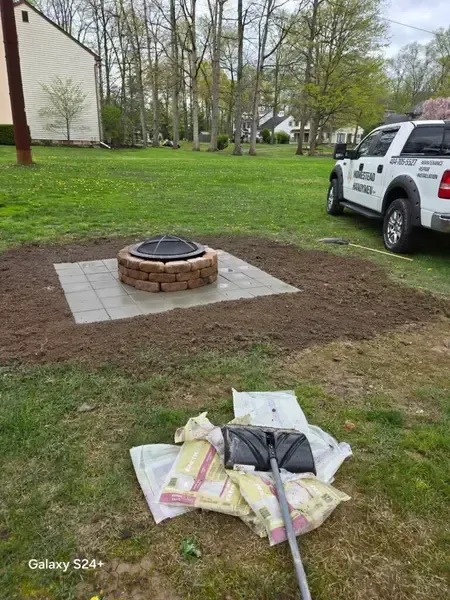 Brick fire pit on a square paver pad in a grassy yard, with landscaping supplies in the foreground.