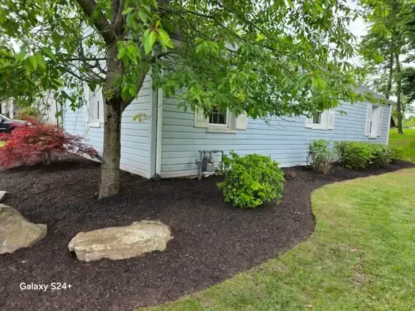 House corner with tree, shrubs, mulch bed, and two large rocks beside a green lawn.