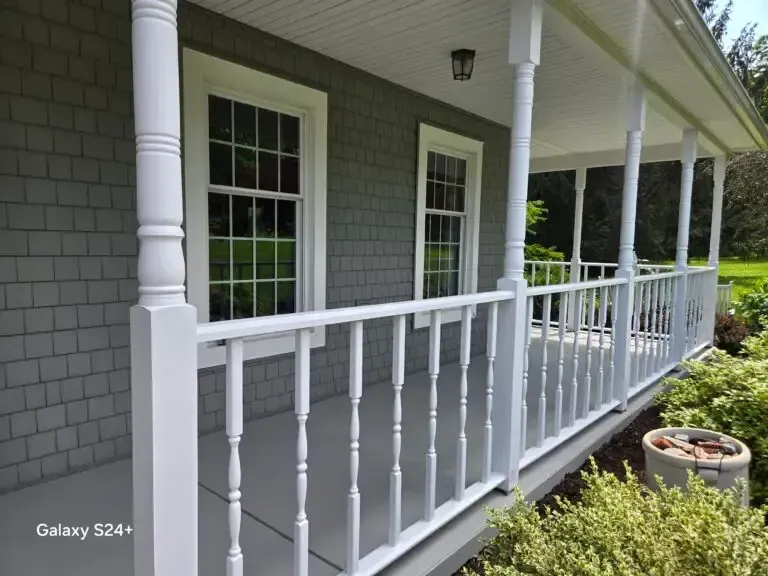 Front porch with white railings and columns on a gray-shingled house, viewed from the side.