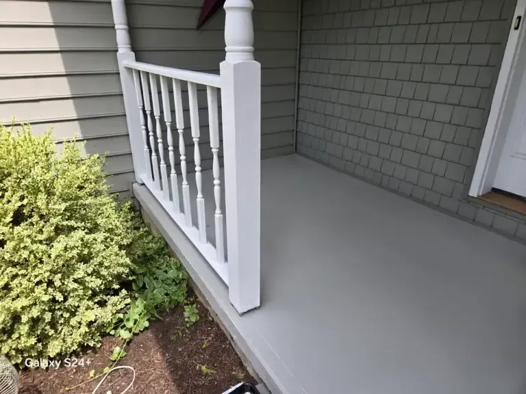 Covered front porch with light gray floor, white railing, and beige siding beside a small shrub.
