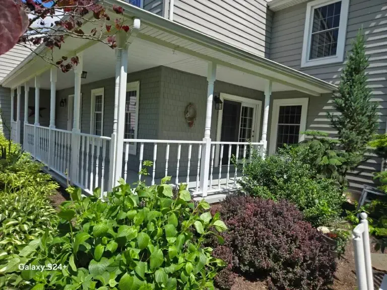 Covered porch with white railing on a beige house, surrounded by green shrubs and flowering plants.