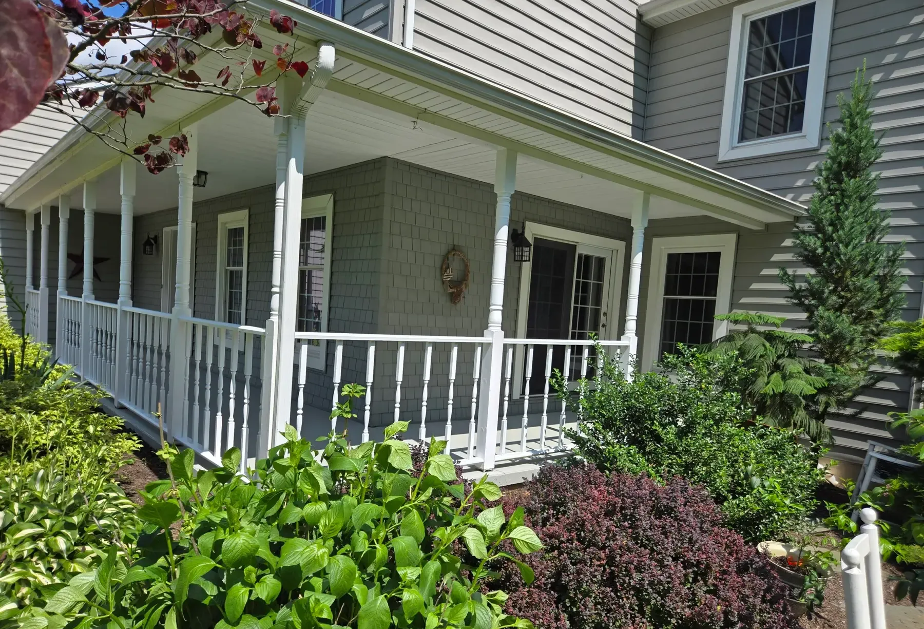 White porch with railing and hanging planter beside a house, surrounded by green shrubs and flowers.
