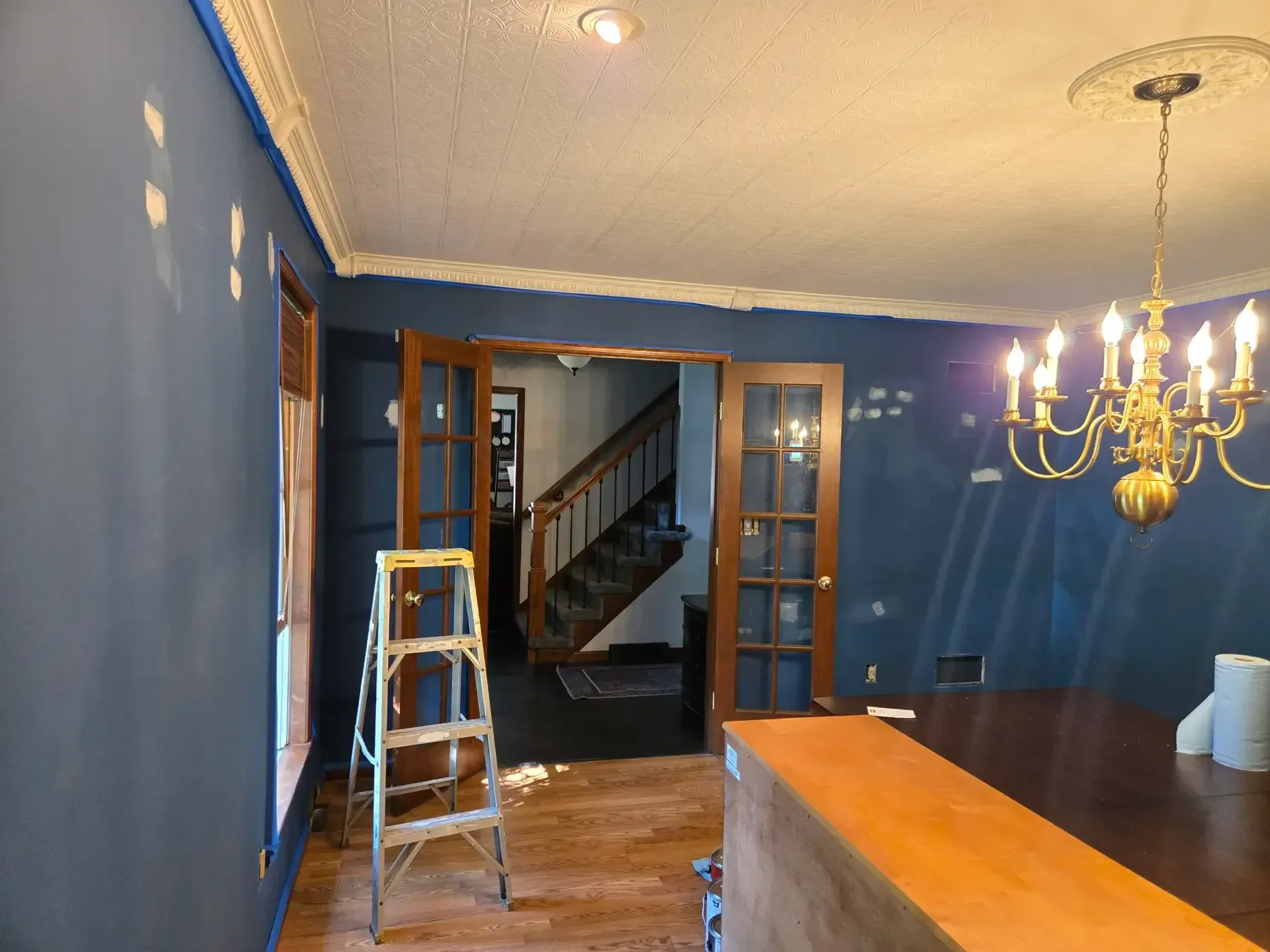 Blue-walled dining room with French doors, a ladder, and a gold chandelier over a dark wood table