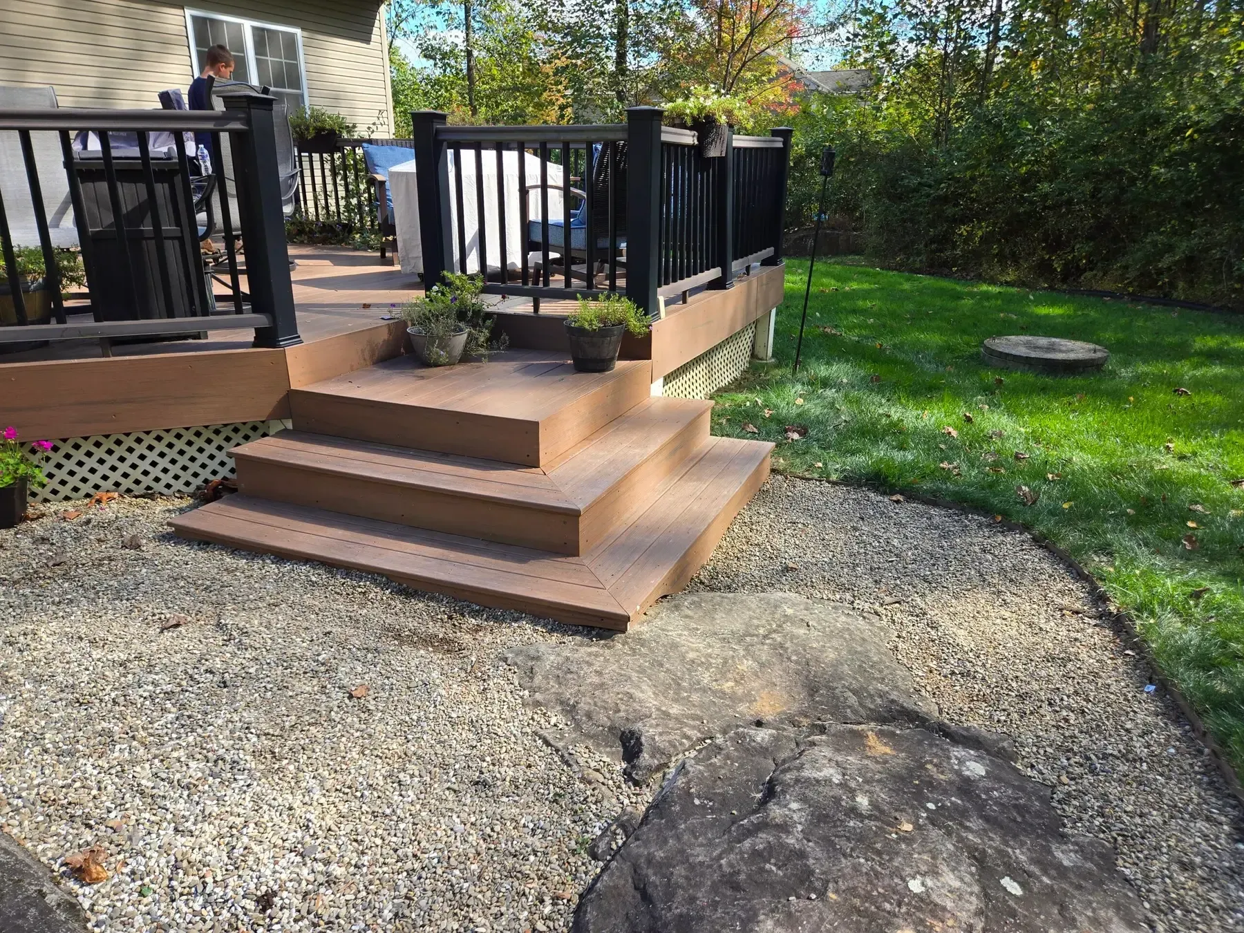 Backyard deck with black railings, tan steps, gravel path, and green lawn bordered by trees.