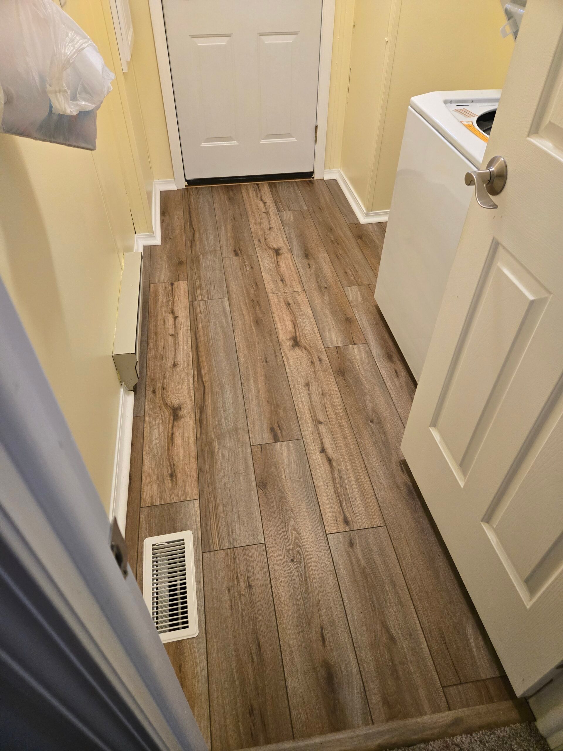 Narrow laundry room hallway with wood-look flooring, open door, and white appliance on the right.