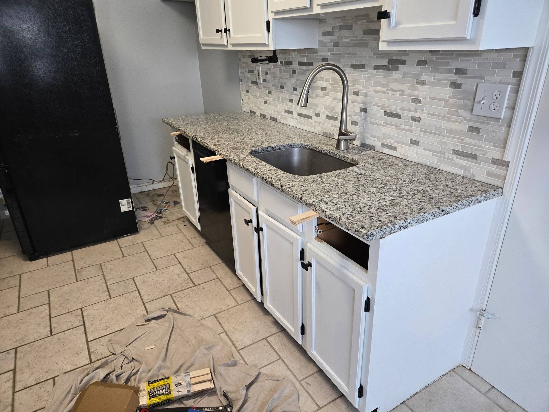 Small kitchen with white cabinets, granite counters, stainless sink, tiled floor, and a black fridge.