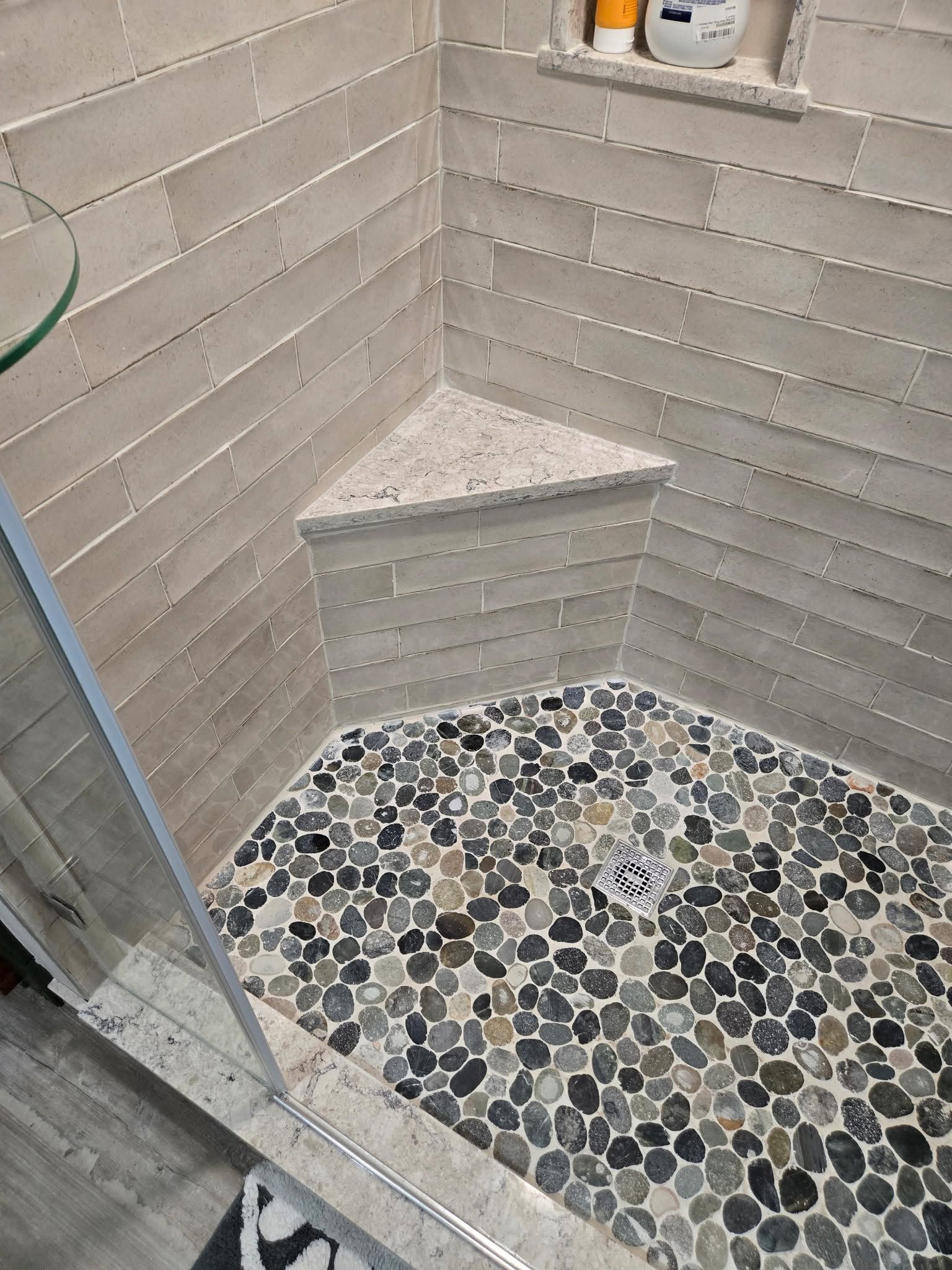 Small tiled shower corner with a pebble floor, white walls, and a built-in triangular shelf.
