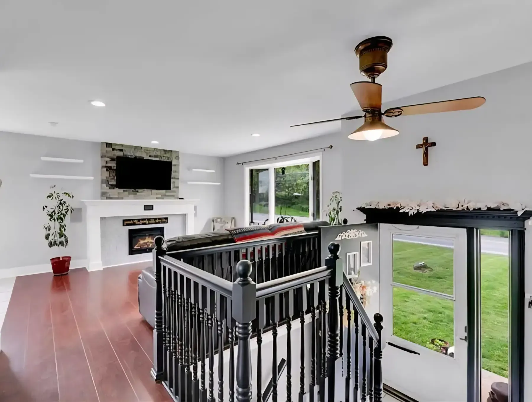 Bright living room with red floor, fireplace, TV, ceiling fan, staircase, and large windows overlooking a green yard