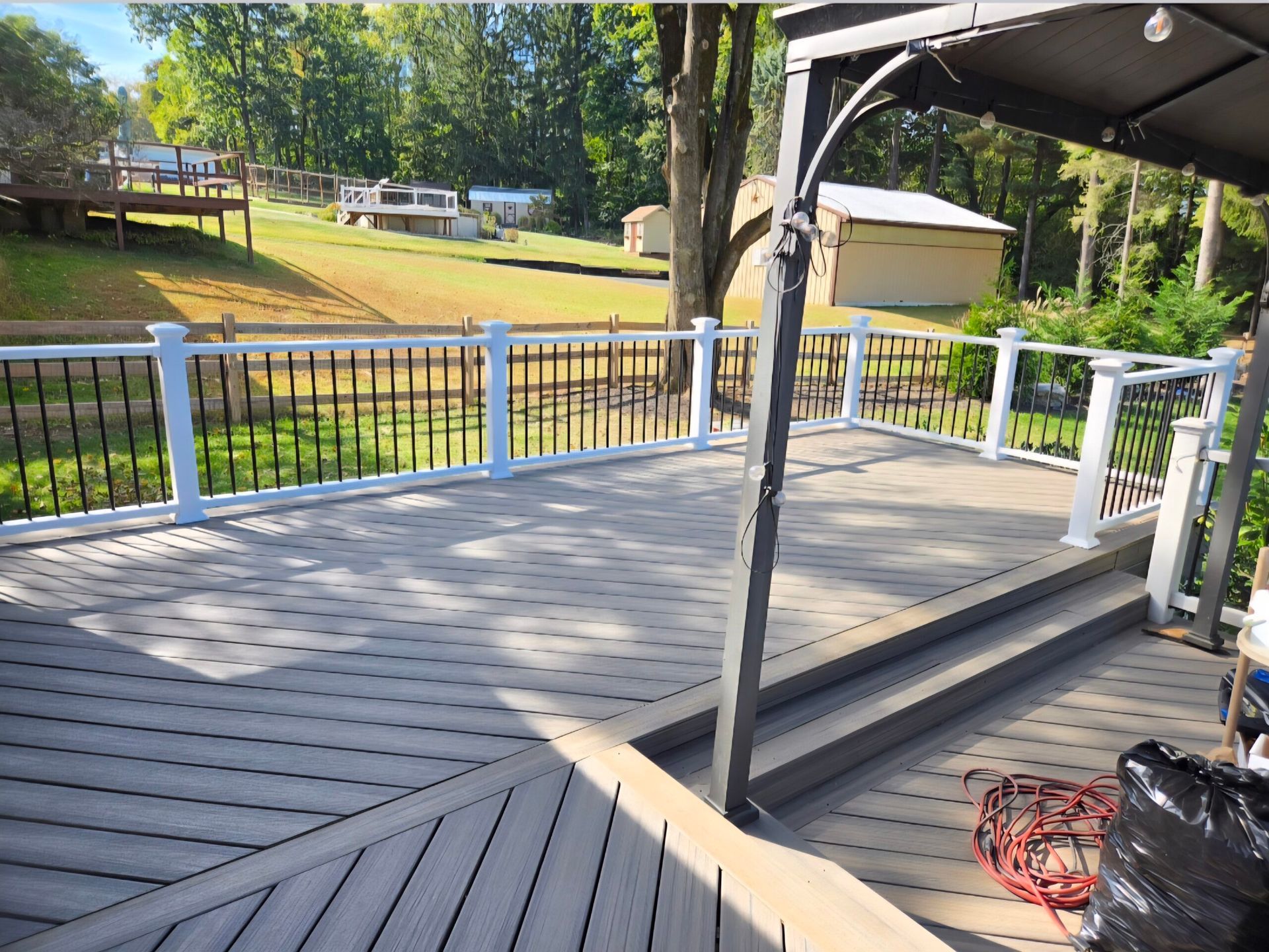 Wooden deck with white railing overlooking a grassy yard and trees, with a canopy overhead and a coiled red hose on the floor