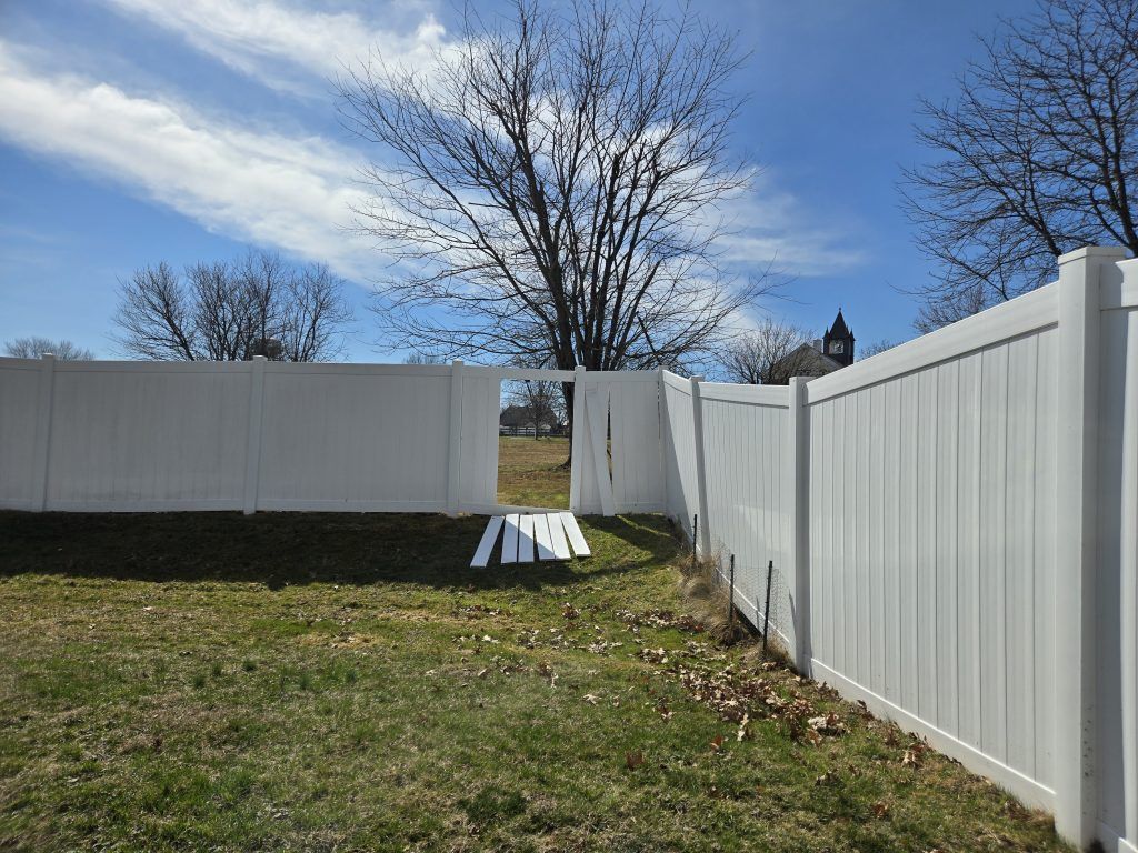 Backyard with white vinyl fence, open gate, and grassy yard under a blue sky
