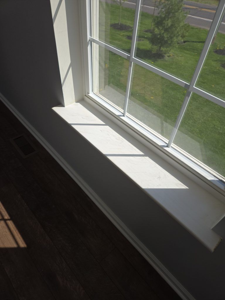 Sunlit white windowsill beside a window, with dark hardwood floor and green lawn outside.